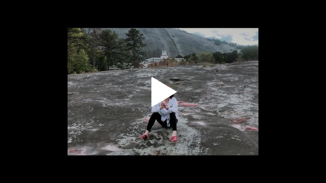 A video still of a woman sitting in the middle of a stream, with mountains and trees behind her