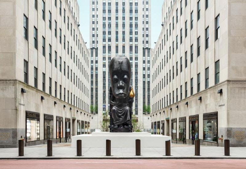 A large black statue of a seated figure with an outsized head in pictured in Rockefeller Plaza